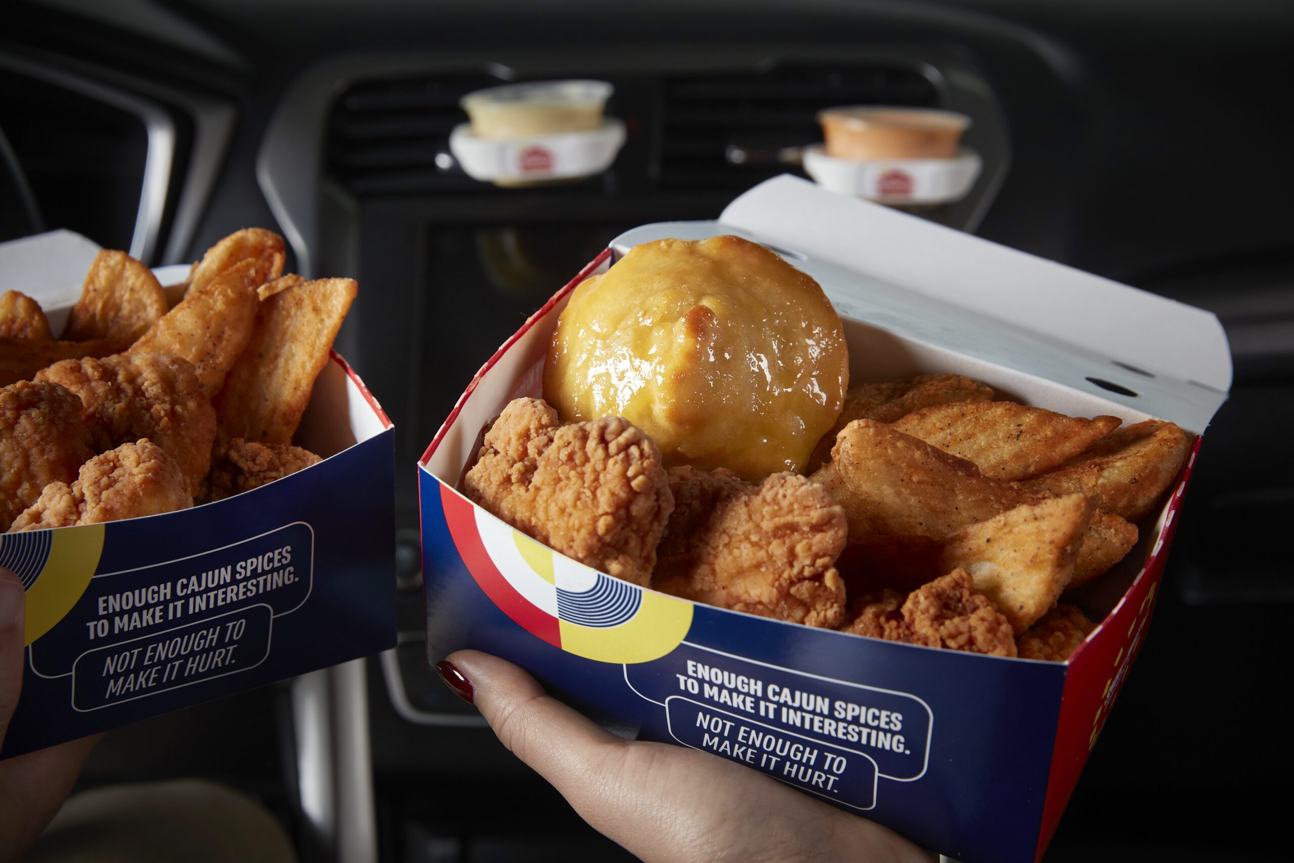 Two Krispy Krunchy Chicken combo boxes with fried chicken, potato wedges, and honey biscuits being held inside a car.