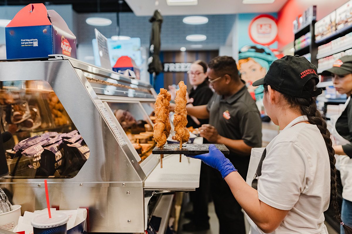Krispy Krunchy Chicken team members preparing fresh fried chicken tenders behind the counter inside a brightly lit convenience store kitchen.
