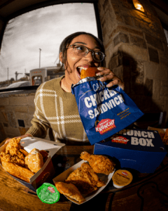 Guest enjoying a Krispy Krunchy Chicken Cajun Chicken Sandwich alongside fried chicken, honey biscuit, wedges, and dipping sauces inside a restaurant.