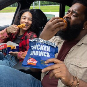 Two guests enjoying Krispy Krunchy Chicken inside a vehicle, with one eating a Cajun Chicken Sandwich and the other holding a crispy chicken tender meal.