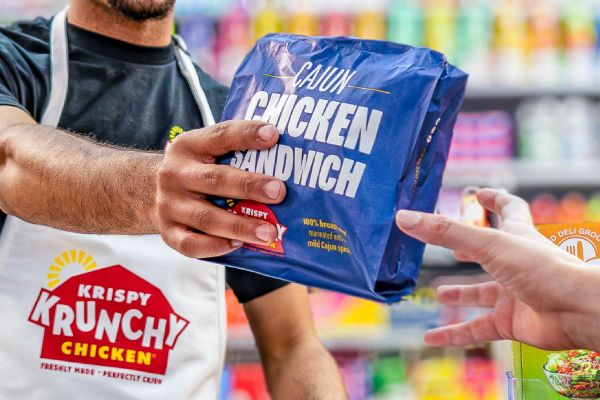 Krispy Krunchy Chicken team member hands a packaged Cajun Chicken Sandwich to a customer in a convenience store