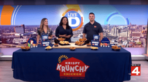 Krispy Krunchy Chicken team member joins WDIV Local 4 hosts in studio with a table full of fried chicken, sides, sauces, and Krunch Box meals.